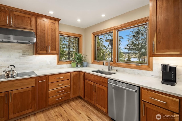 kitchen featuring brown cabinetry, stainless steel dishwasher, a sink, light countertops, and under cabinet range hood
