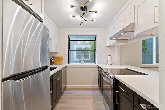 kitchen with appliances with stainless steel finishes, a chandelier, white cabinetry, light wood-type flooring, and under cabinet range hood