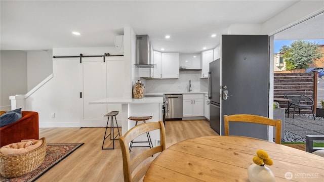 kitchen with appliances with stainless steel finishes, a barn door, light countertops, light wood-style floors, and wall chimney exhaust hood
