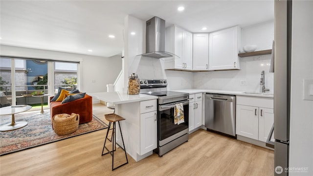kitchen featuring wall chimney range hood, a breakfast bar, a sink, stainless steel appliances, and light wood-type flooring