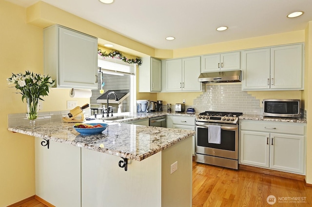 kitchen featuring light stone countertops, a sink, a peninsula, under cabinet range hood, and stainless steel appliances