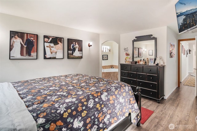 bedroom featuring arched walkways, baseboards, ensuite bath, and wood finished floors