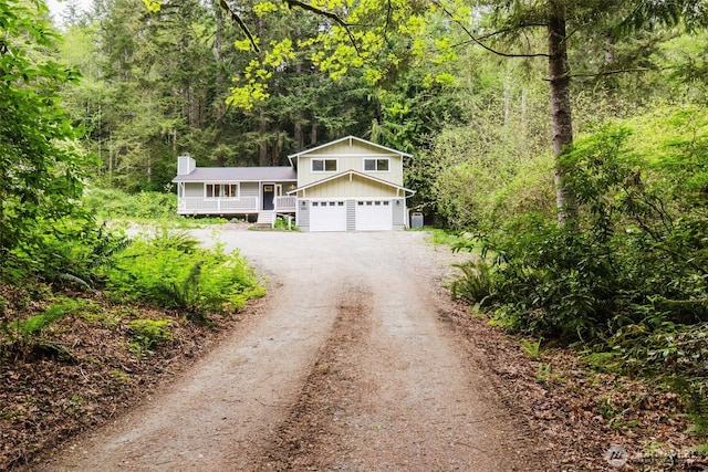 split level home featuring board and batten siding, a forest view, a chimney, dirt driveway, and an attached garage
