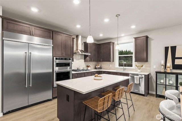 kitchen with wall chimney range hood, dark brown cabinets, a sink, light wood-style floors, and stainless steel appliances