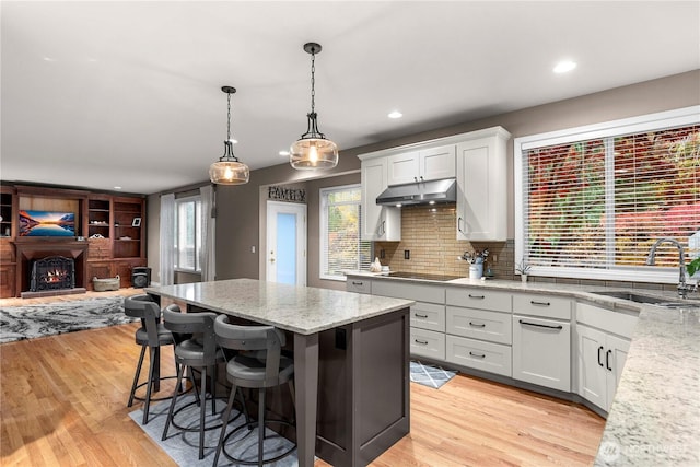 kitchen with a warm lit fireplace, black electric stovetop, a sink, light wood finished floors, and under cabinet range hood
