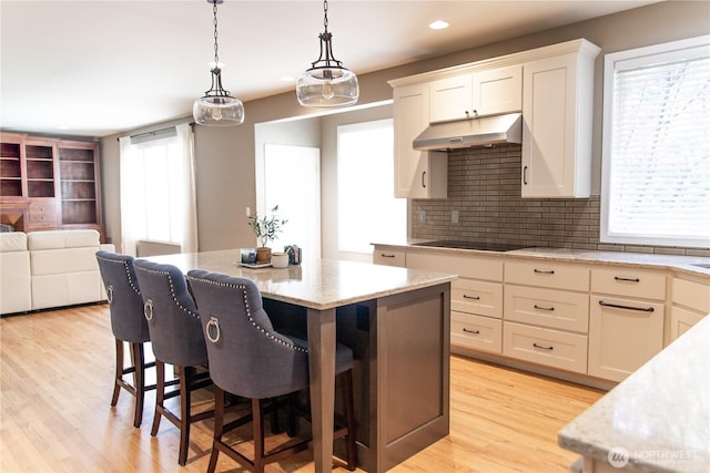 kitchen with light stone countertops, a kitchen breakfast bar, black electric stovetop, under cabinet range hood, and tasteful backsplash