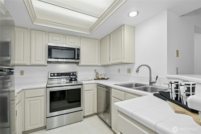 kitchen with a tray ceiling, a sink, light tile patterned floors, tile counters, and stainless steel appliances