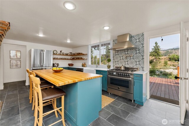 kitchen with wall chimney range hood, blue cabinets, open shelves, butcher block counters, and stainless steel appliances