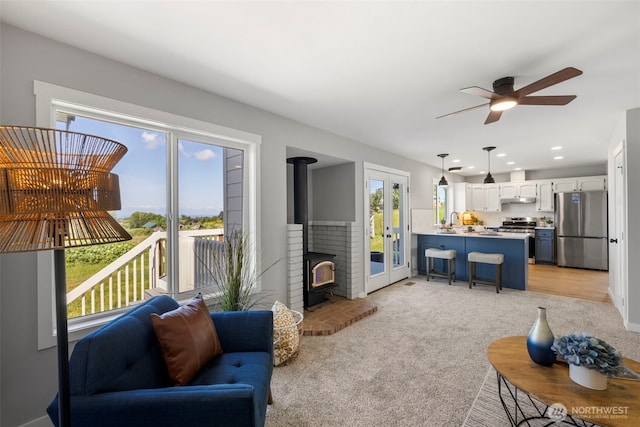 living area with a wood stove, ceiling fan, light colored carpet, and french doors