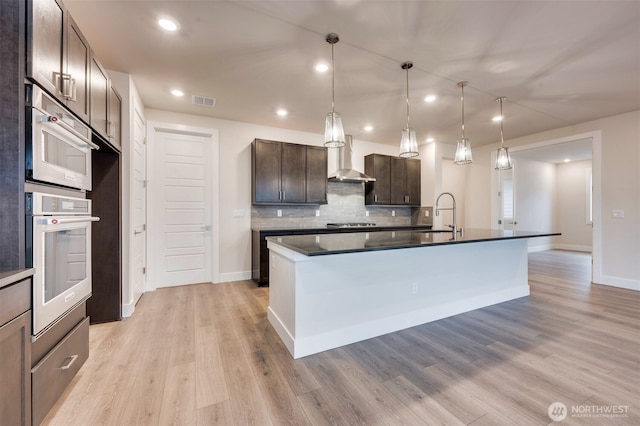 kitchen featuring wall chimney range hood, dark countertops, dark brown cabinetry, light wood-style floors, and decorative backsplash