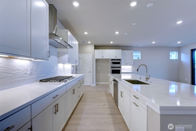 kitchen featuring wall chimney range hood, appliances with stainless steel finishes, a sink, white cabinetry, and tasteful backsplash