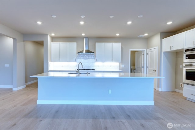 kitchen with white cabinets, an island with sink, light wood-style flooring, wall chimney exhaust hood, and backsplash