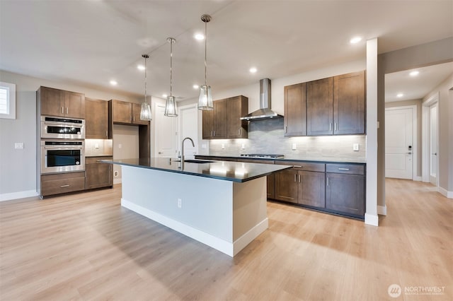 kitchen with backsplash, dark countertops, wall chimney exhaust hood, a sink, and light wood-style flooring