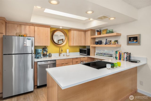 kitchen featuring a peninsula, light brown cabinetry, a tray ceiling, a sink, and appliances with stainless steel finishes