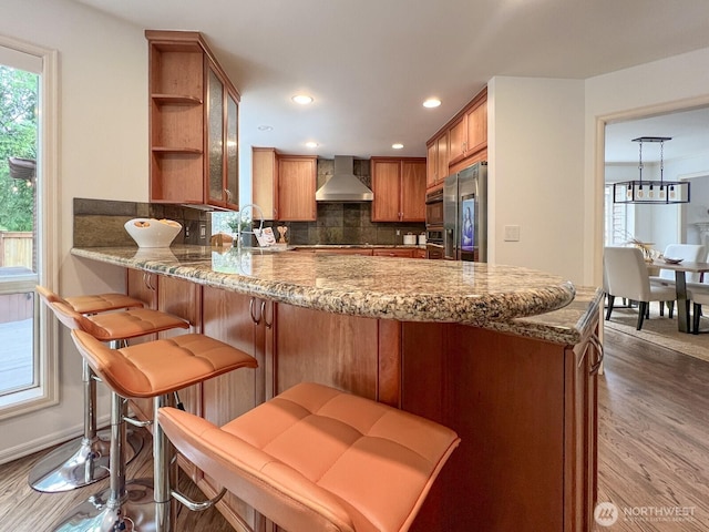 kitchen with decorative backsplash, brown cabinets, a peninsula, stainless steel fridge, and wall chimney exhaust hood