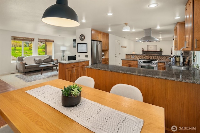 kitchen with brown cabinetry, stainless steel appliances, open floor plan, a peninsula, and range hood