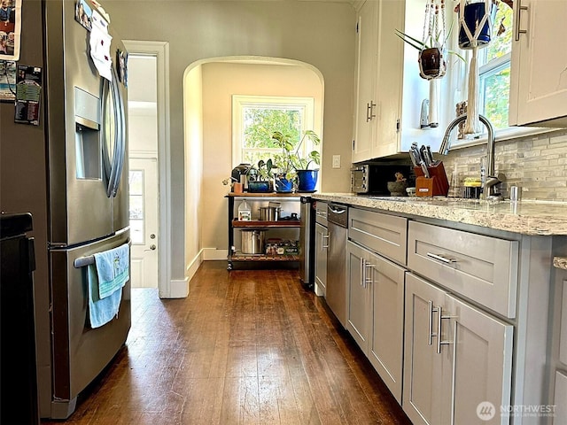 kitchen featuring stainless steel appliances, arched walkways, backsplash, dark wood-style floors, and light stone countertops