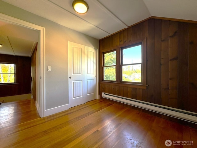 unfurnished bedroom with wooden walls, wood finished floors, baseboard heating, multiple windows, and vaulted ceiling