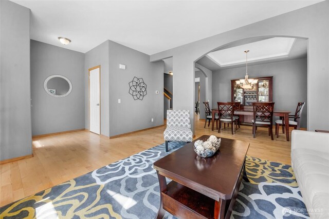 living room featuring arched walkways, baseboards, a raised ceiling, a chandelier, and light wood-style floors