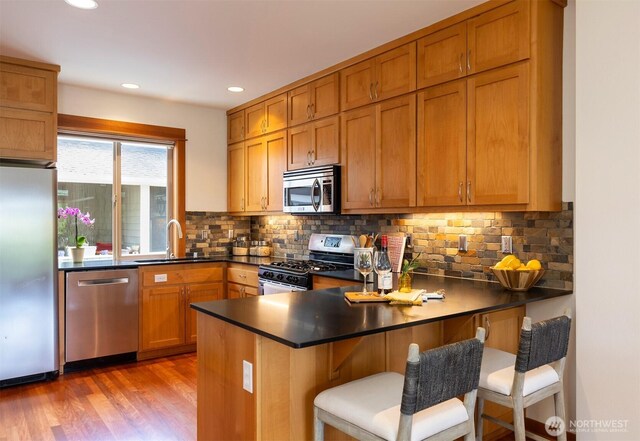 kitchen featuring dark countertops, appliances with stainless steel finishes, a sink, brown cabinetry, and a peninsula