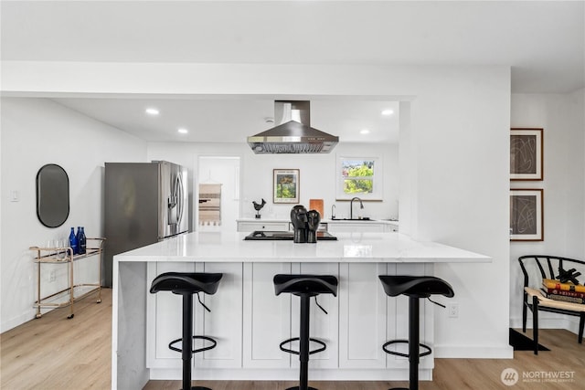 kitchen featuring wall chimney range hood, light wood-style floors, a kitchen breakfast bar, a peninsula, and cooktop
