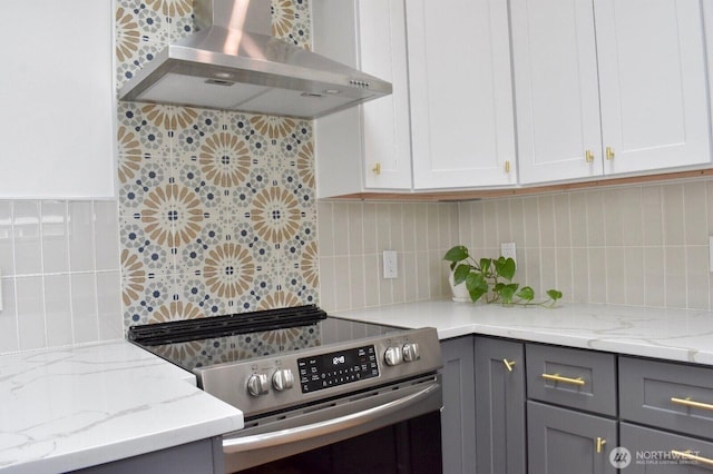 kitchen with gray cabinets, wall chimney exhaust hood, light stone counters, electric stove, and backsplash