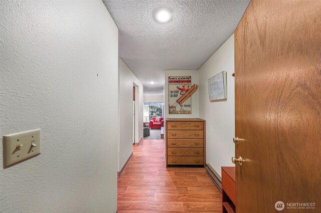 hallway featuring a textured ceiling, baseboard heating, baseboards, light wood finished floors, and a textured wall