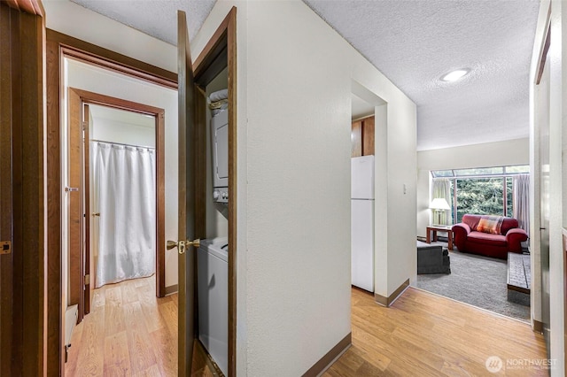 hallway with a textured ceiling, baseboards, stacked washer and clothes dryer, and light wood-type flooring