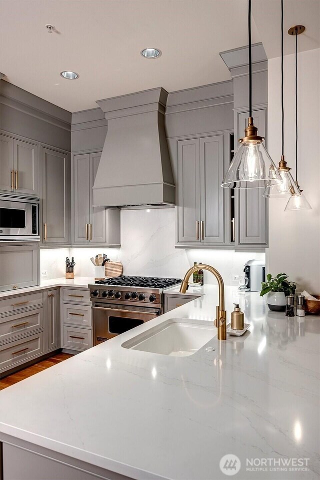 kitchen with light stone countertops, gray cabinets, custom range hood, a sink, and appliances with stainless steel finishes