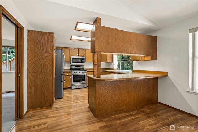 kitchen with brown cabinetry, appliances with stainless steel finishes, vaulted ceiling, a peninsula, and light wood finished floors