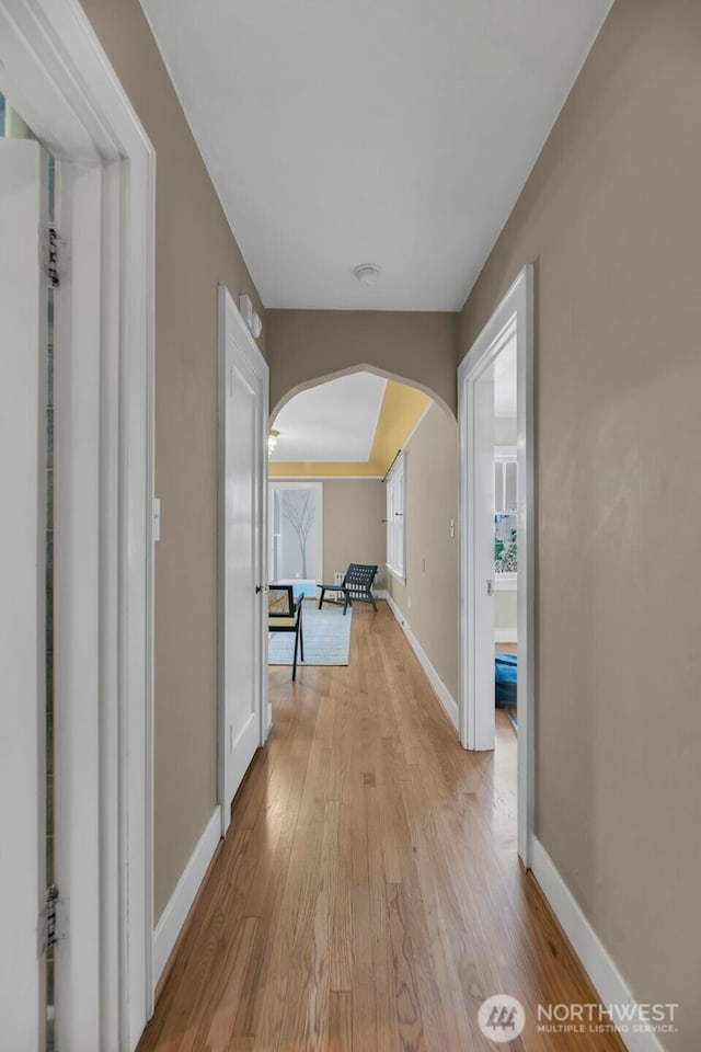 corridor featuring arched walkways, light wood-type flooring, and baseboards