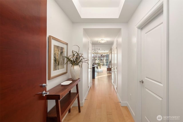 hall with baseboards, a raised ceiling, and light wood finished floors