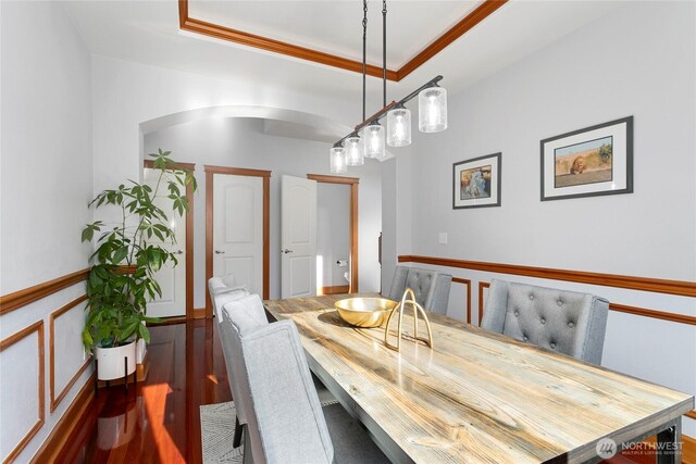 dining room with wood finished floors, arched walkways, ornamental molding, a raised ceiling, and wainscoting