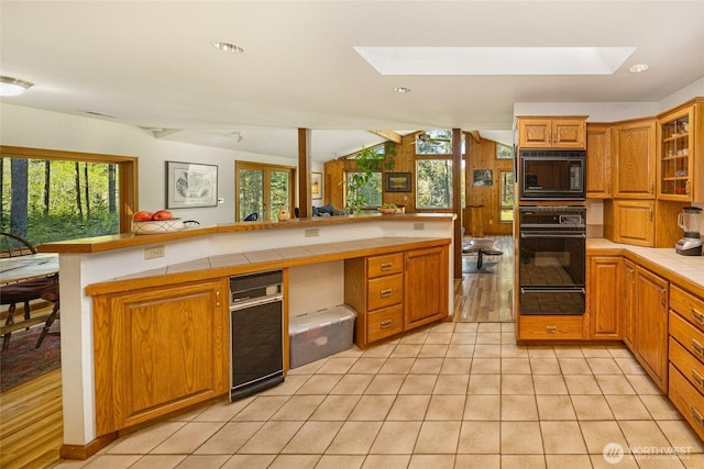 kitchen with glass insert cabinets, lofted ceiling with skylight, tile countertops, a warming drawer, and black appliances
