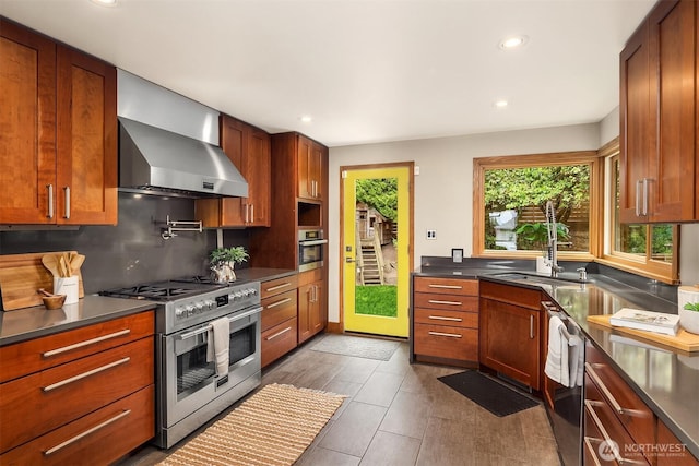 kitchen featuring appliances with stainless steel finishes, wall chimney range hood, dark countertops, decorative backsplash, and recessed lighting