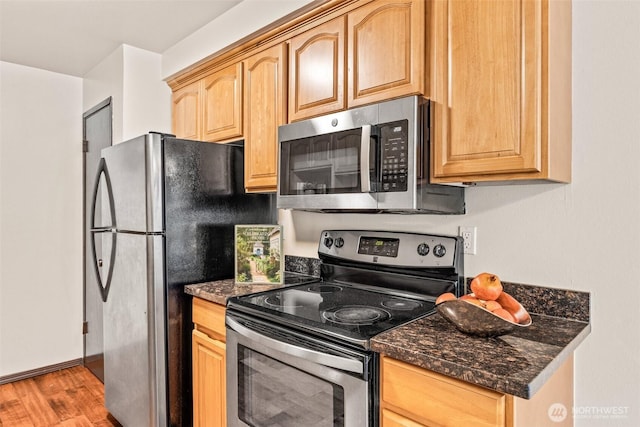 kitchen with stainless steel appliances, light brown cabinetry, light wood-type flooring, dark stone countertops, and baseboards