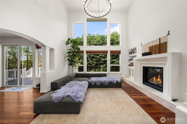 living area with an inviting chandelier, a glass covered fireplace, a towering ceiling, wood finished floors, and arched walkways