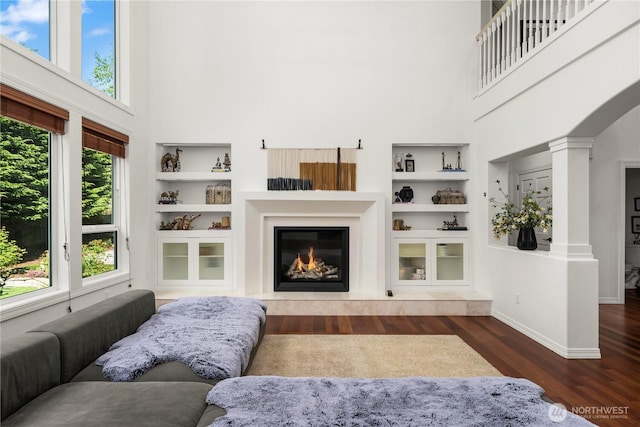 living area featuring built in features, wood finished floors, baseboards, a glass covered fireplace, and a high ceiling