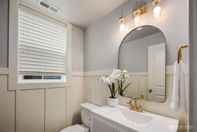 half bathroom with visible vents, vanity, wainscoting, a decorative wall, and toilet