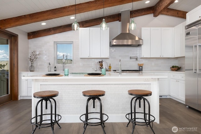 kitchen featuring light countertops, an island with sink, wall chimney exhaust hood, vaulted ceiling with beams, and stainless steel built in refrigerator