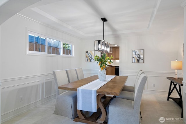 dining area featuring a raised ceiling, a decorative wall, light colored carpet, arched walkways, and wainscoting