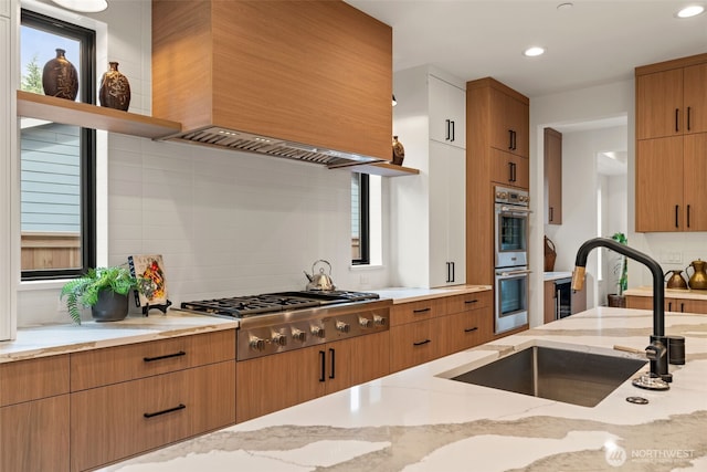 kitchen featuring decorative backsplash, appliances with stainless steel finishes, light stone countertops, a sink, and premium range hood