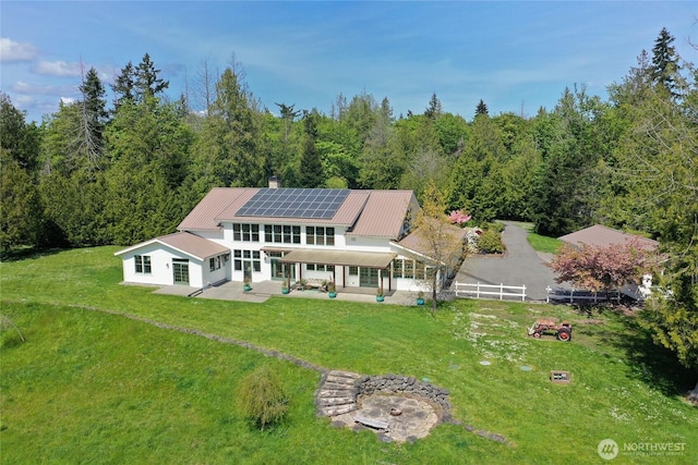 rear view of house featuring a patio, a view of trees, a lawn, and solar panels