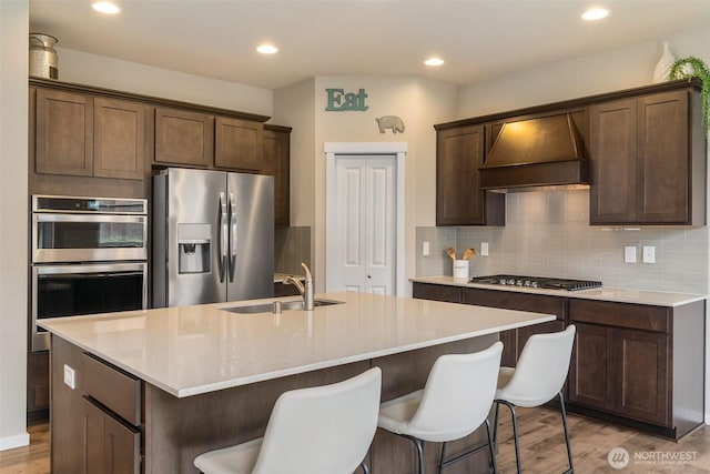 kitchen featuring a center island with sink, light wood-type flooring, a sink, custom exhaust hood, and stainless steel appliances