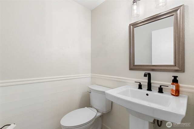 half bathroom with toilet, tile walls, a sink, and a wainscoted wall