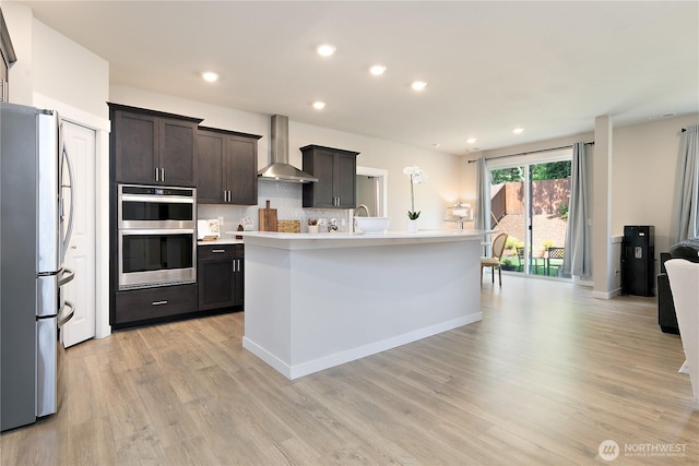 kitchen with wall chimney range hood, appliances with stainless steel finishes, light countertops, decorative backsplash, and light wood-type flooring
