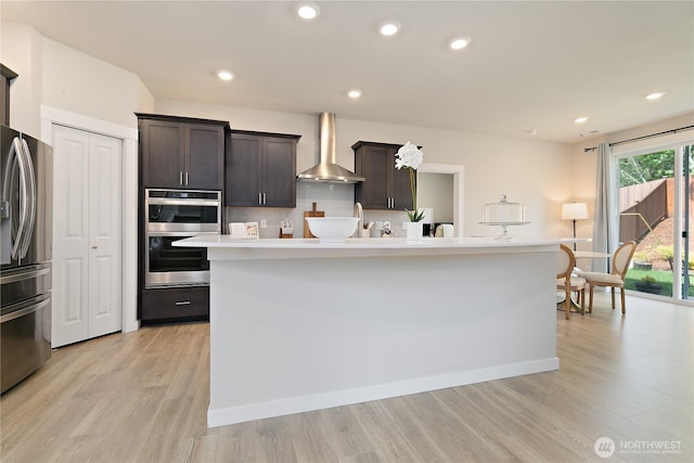 kitchen featuring appliances with stainless steel finishes, light wood-style flooring, wall chimney exhaust hood, and dark brown cabinetry