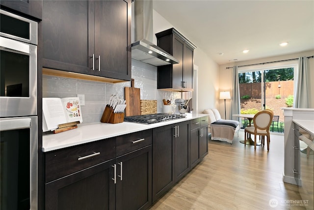 kitchen with light countertops, light wood-style flooring, wall chimney exhaust hood, stainless steel appliances, and backsplash