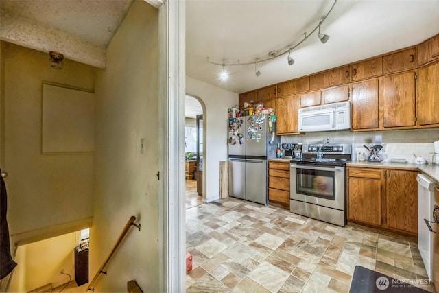kitchen featuring stone finish floor, arched walkways, brown cabinets, light countertops, and stainless steel appliances