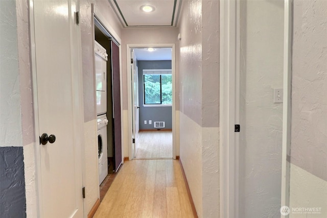 hall featuring light wood-style floors, a textured wall, stacked washer / dryer, and visible vents